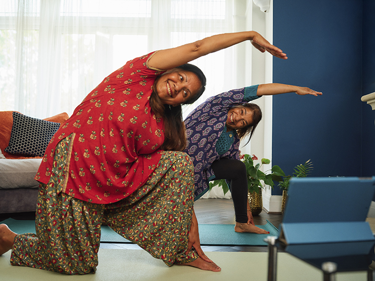 Two women perform yoga at home, following a class on a screen in front of them