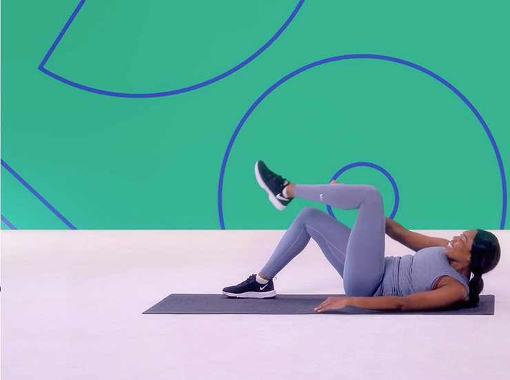 A woman lays on an exercise mat in a studio, with a green background, and performs crunches to the opposing knee