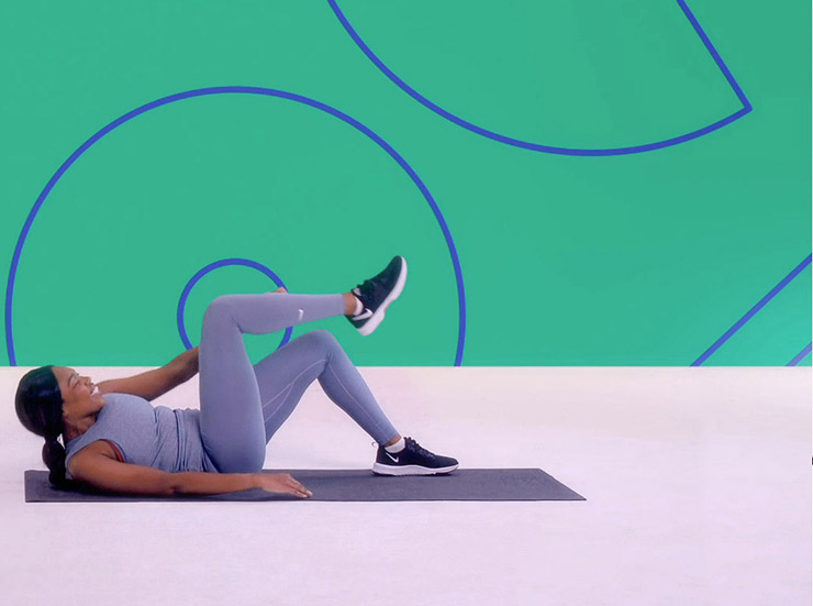 A woman lays on an exercise mat in a studio, with a green background, and performs crunches to the opposing knee