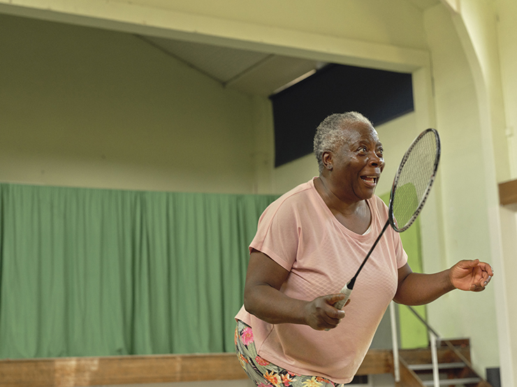 An older black woman with short grey hair stands ready to receive a serve in a game of badminton