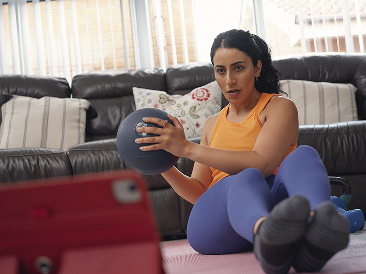 A woman uses a medicine ball to perform Russian twists during a home exercise session, while watching an instructor on an iPad