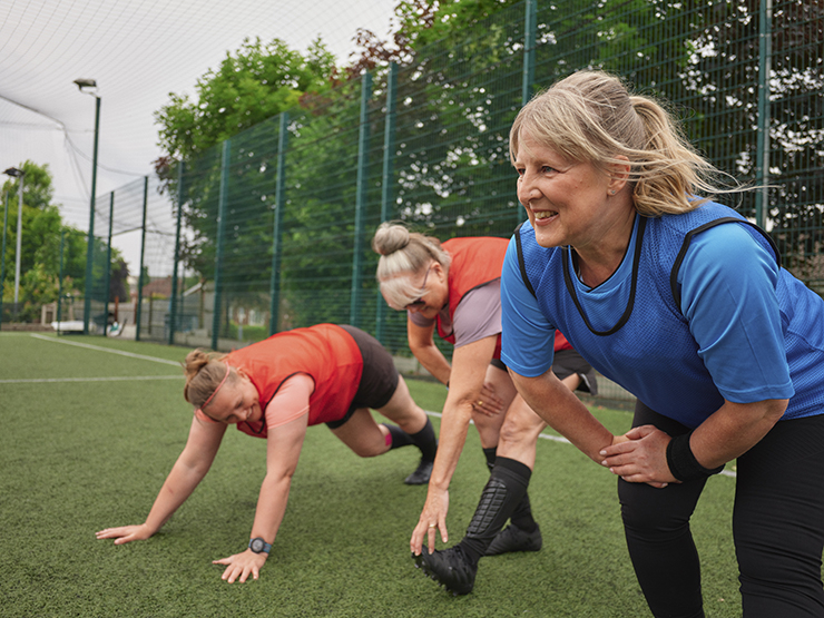 Three older women prepare for a walking football match with leg stretches on the pitch