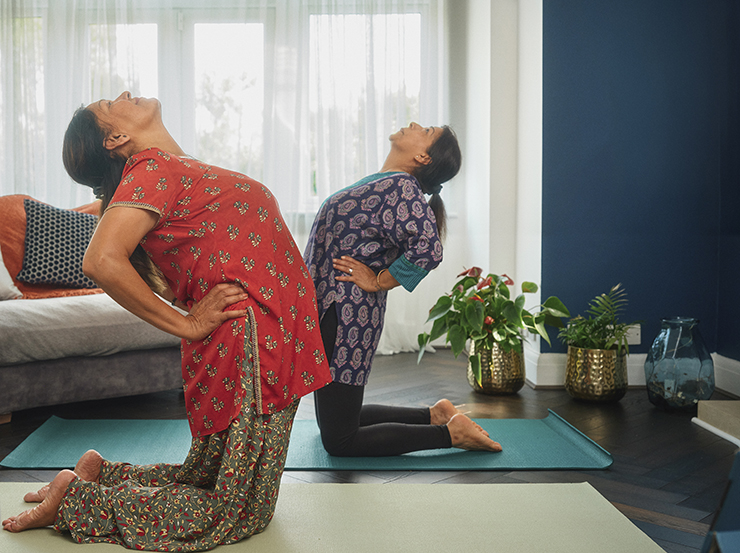 Two South Asian women perform yoga at home. Both face each other, one closer to the camera than the other, on their knees, while arching their backs and looking to the ceiling, with hands on their hips.