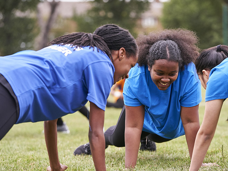 Three young girls adopt a press-up position on the grass outside, all facing each other in a circle