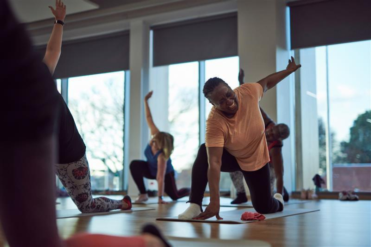 A woman stretches her arm up while on one knee on a yoga mat in a class, alongside other participants.