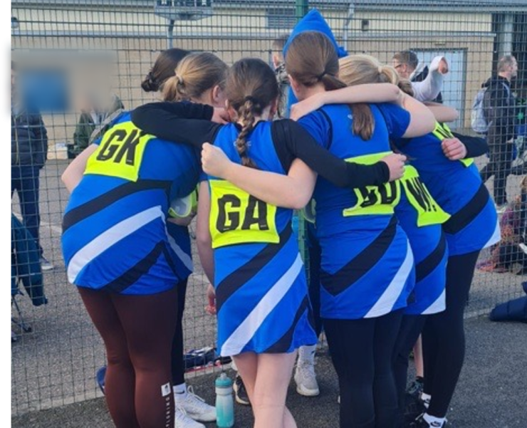 A girls' netball team huddle in a circle with their arms around each other.