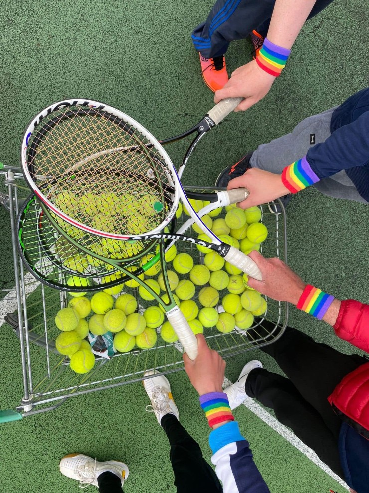 Aerial view of several arms holding tennis racquets over a trolley of balls on a court, with some arms sporting rainbow wristbands.