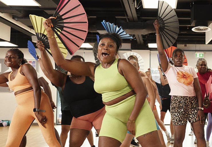 A group of women dance on an indoors studio carrying colourful fans on their hands.