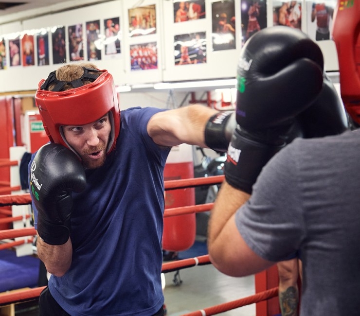 A male boxer wearing a headguard punches pads being held by his sparring partner in a ring.