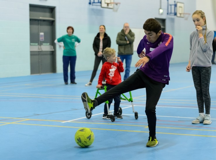 A disabled boy kicks a football in a sports hall as fellow players and parents watch on.