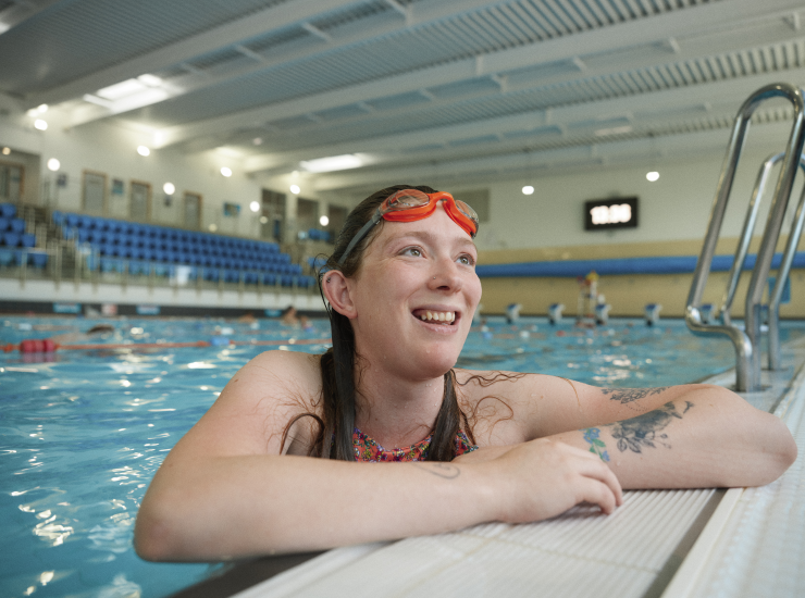 A woman in swimwear and goggles smiles as she leans on the side of a public swimming pool.