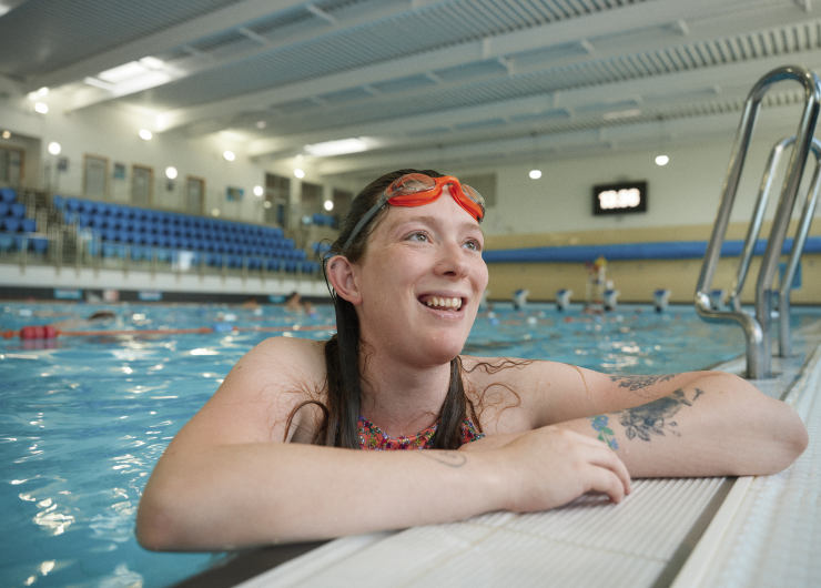 A woman in swimwear and goggles smiles as she leans on the side of a public swimming pool.