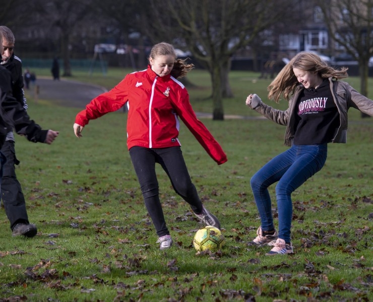 Two girls compete for a football during a casual game on an autumnal-looking park, while two fellow players look on.