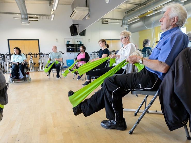 A group of older adults sit on chairs in an exercise studio and perform seated stretching exercises using green resistance bands
