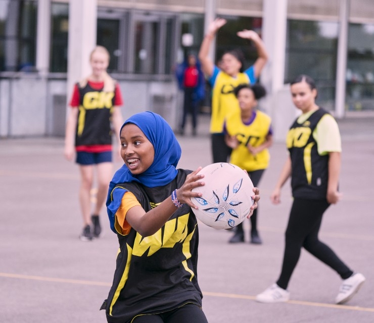 A girl wearing a head scarf takes possession of a netball on a school playground, as other players look on,