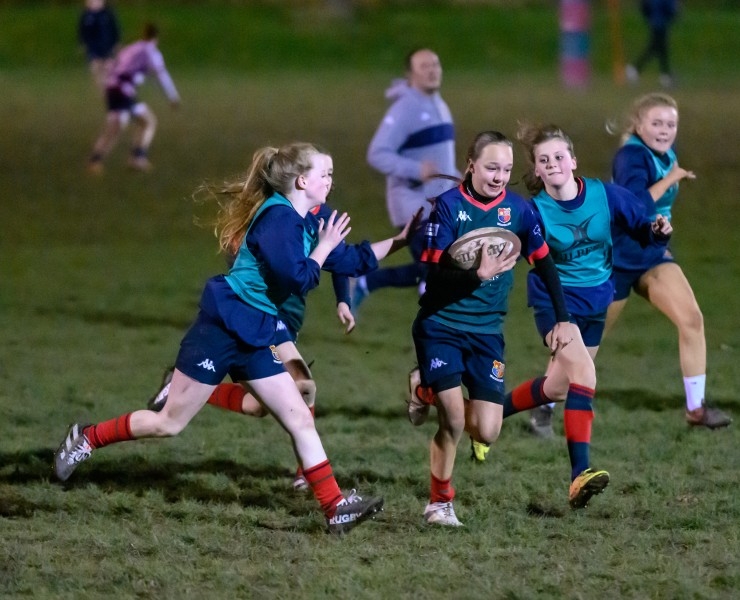 A young female rugby player runs with the ball as opposing players attempt to tackle her in a training game at night.