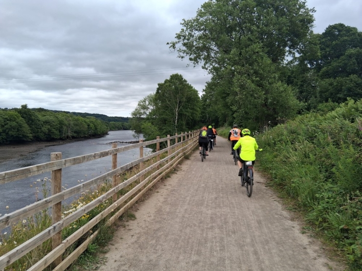 Four cyclists ride on a tree-lined path beside a river.