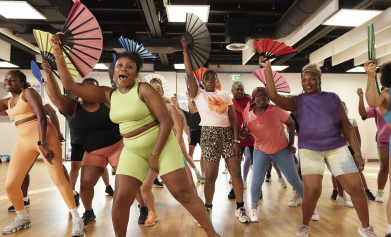 A group of women perform a dance routine in a class, holding and waving paper fans